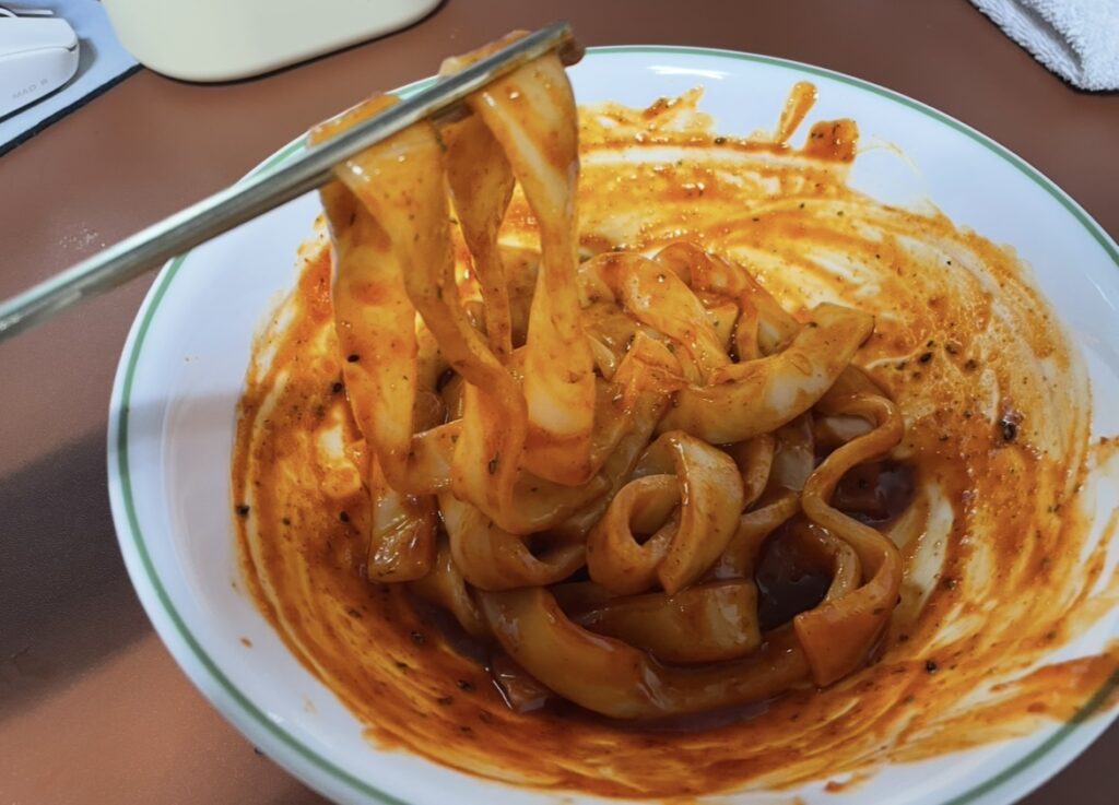 Close-up of cooked flat tteokbokki noodles lifted with chopsticks, coated in thick sauce that highlights Spicy Korean snack flavors.