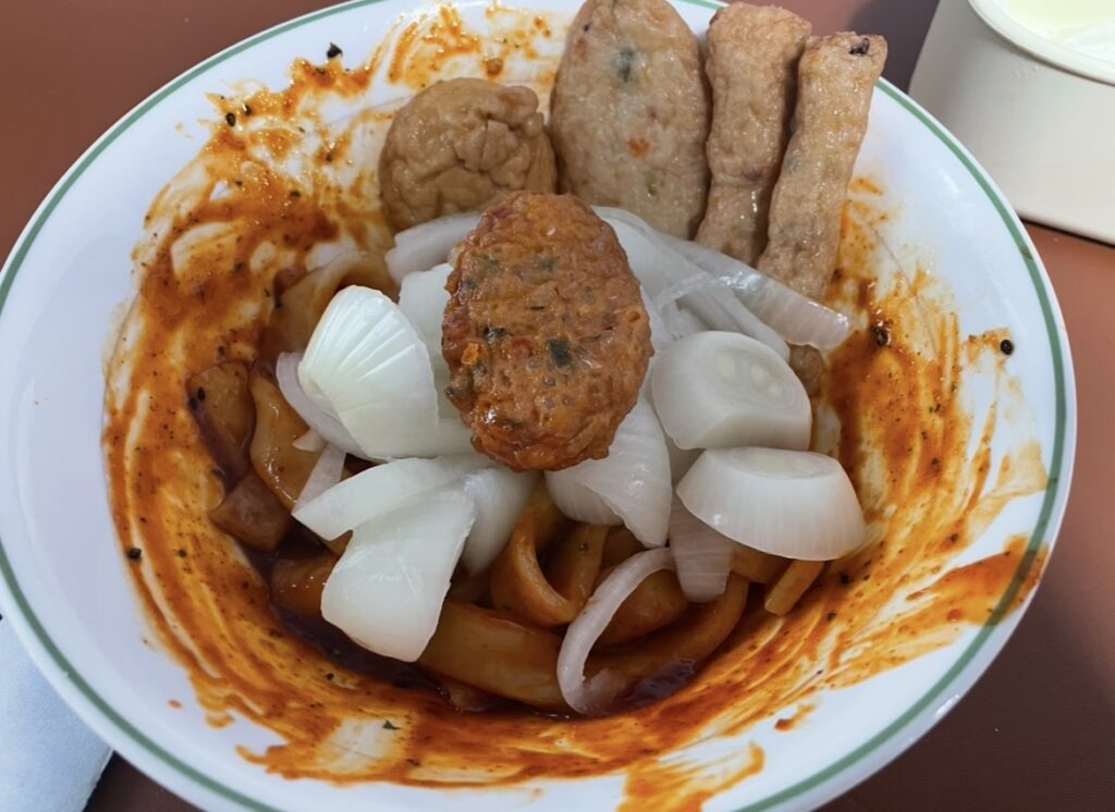 Seasoned tteokbokki noodles topped with fish cakes and onions, a homemade style often seen in a Korean snack review.