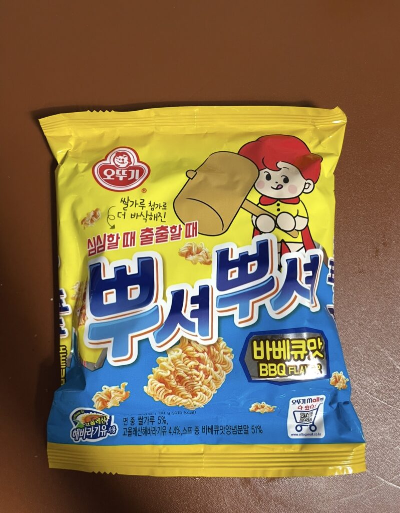 the front of the bbusyeo bbusyeo barbecue ramen snack package shows a bright yellow and blue contrast with a cute character holding a hammer.