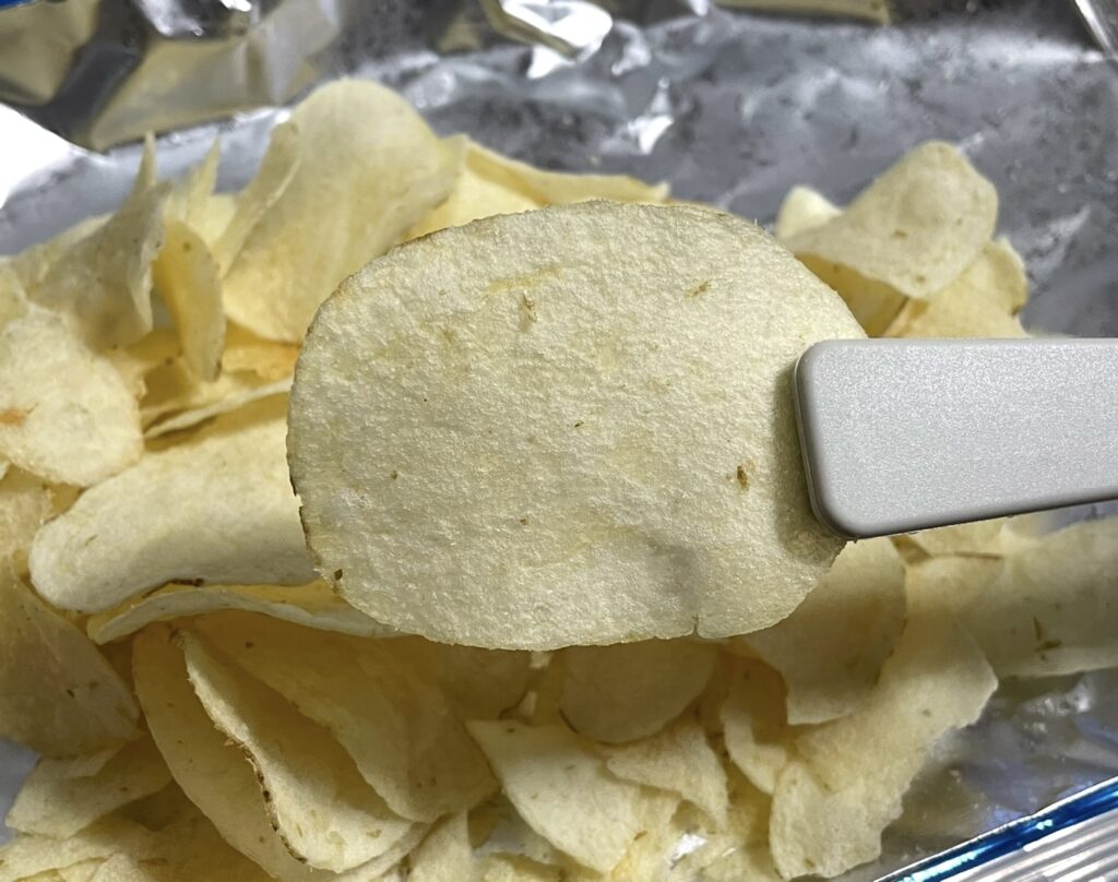A single thin potato chip held with tongs, highlighting its golden color and crisp texture. This close-up is ideal for a Korean snack review to showcase the product’s real appearance
